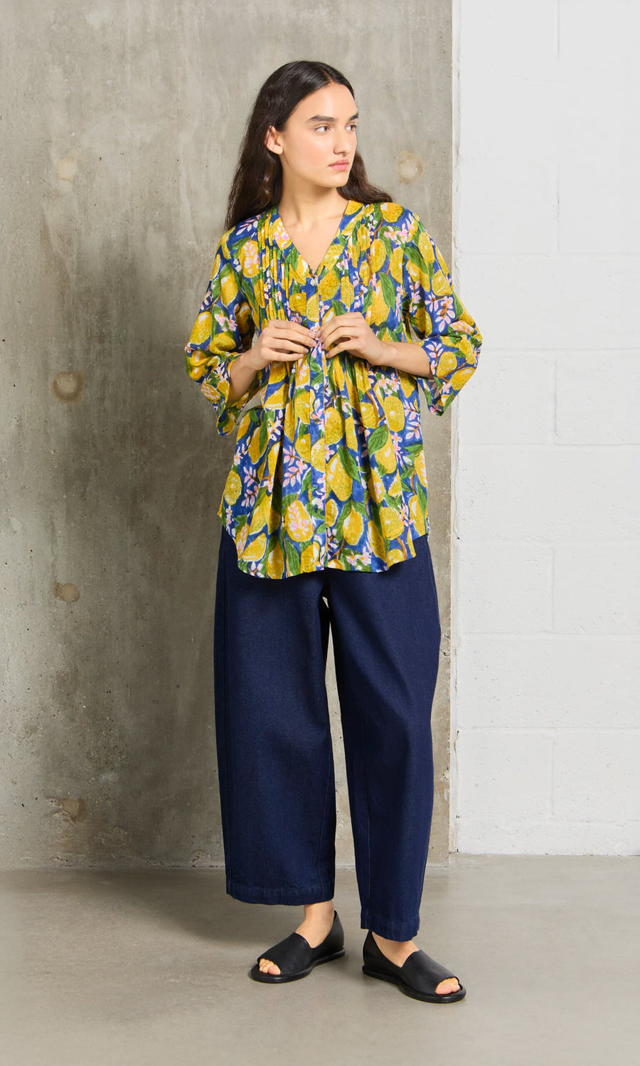 Plümo- A person in a Citrus Shirt, wide-leg jeans, and black sandals stands indoors by a concrete wall.