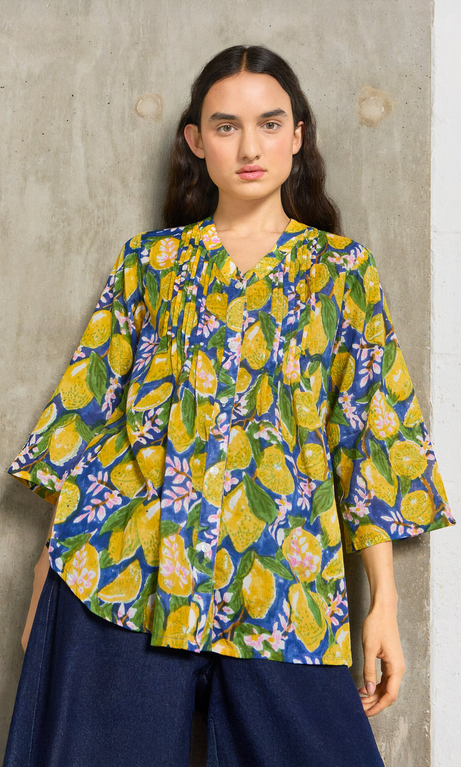 Plümo- Woman in the Citrus Shirt, a loose blouse with yellow and green leaves, stands against a concrete wall.