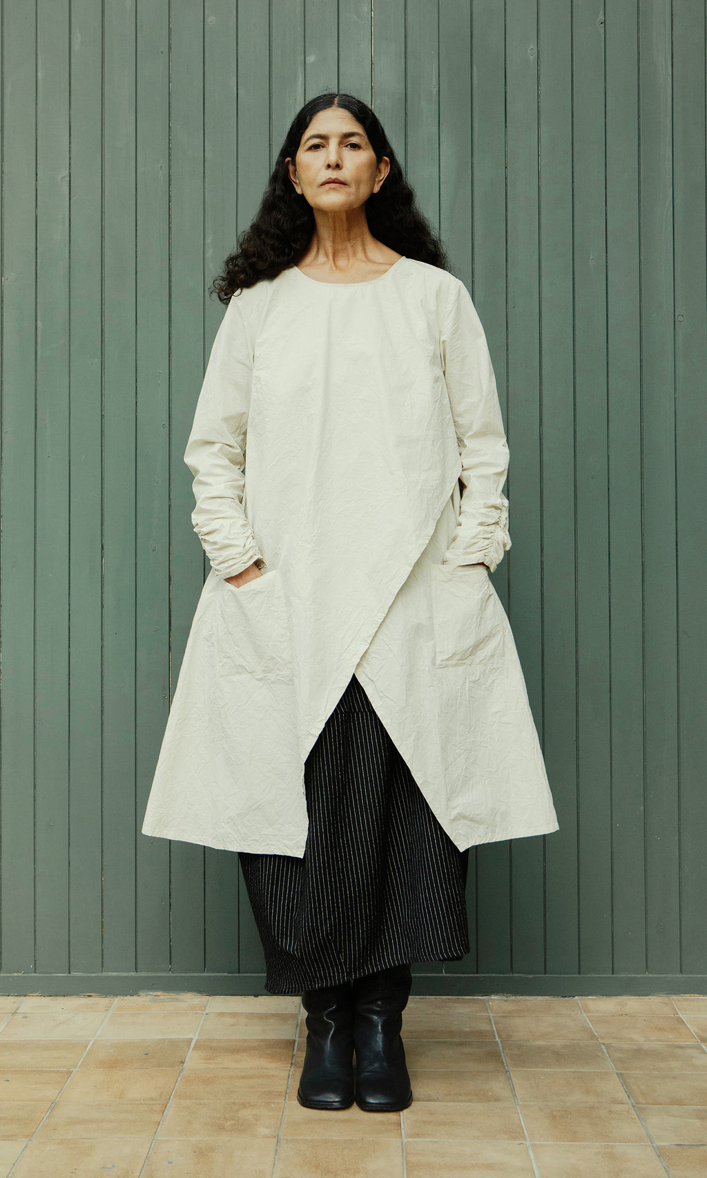 Plümo  Woman with long dark hair in Clementine Blouse, styled with Swedish design, against a green wooden wall.