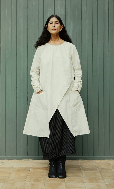 Plümo  Woman with long dark hair in Clementine Blouse, styled with Swedish design, against a green wooden wall.