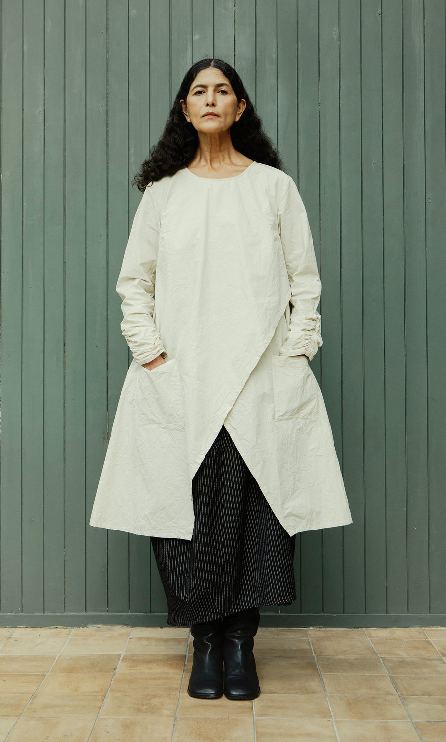 Plümo  Woman with long dark hair in Clementine Blouse, styled with Swedish design, against a green wooden wall.