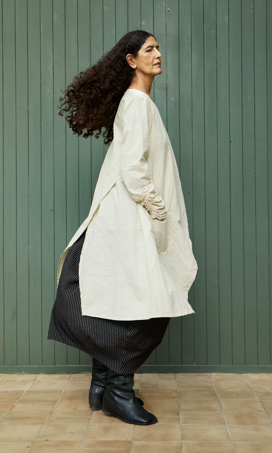 Plümo  Woman with long curly hair wears the Clementine Blouse, a striped skirt, and black boots by a green wall.