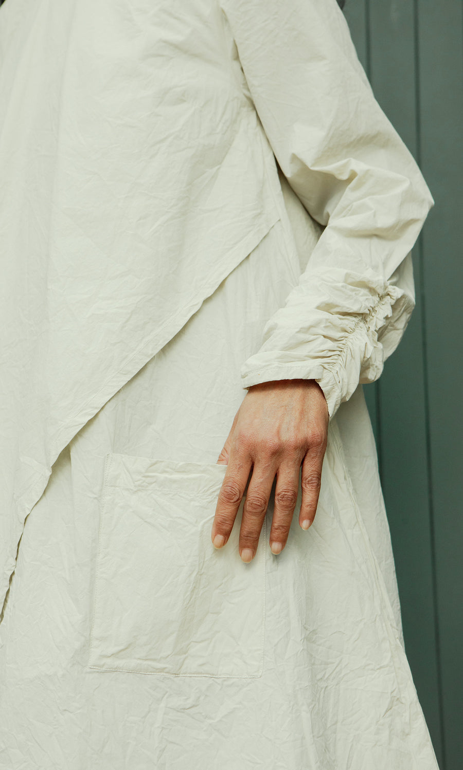 Plümo  Someone in the Clementine Blouse stands with a hand by a pocket against a green background.