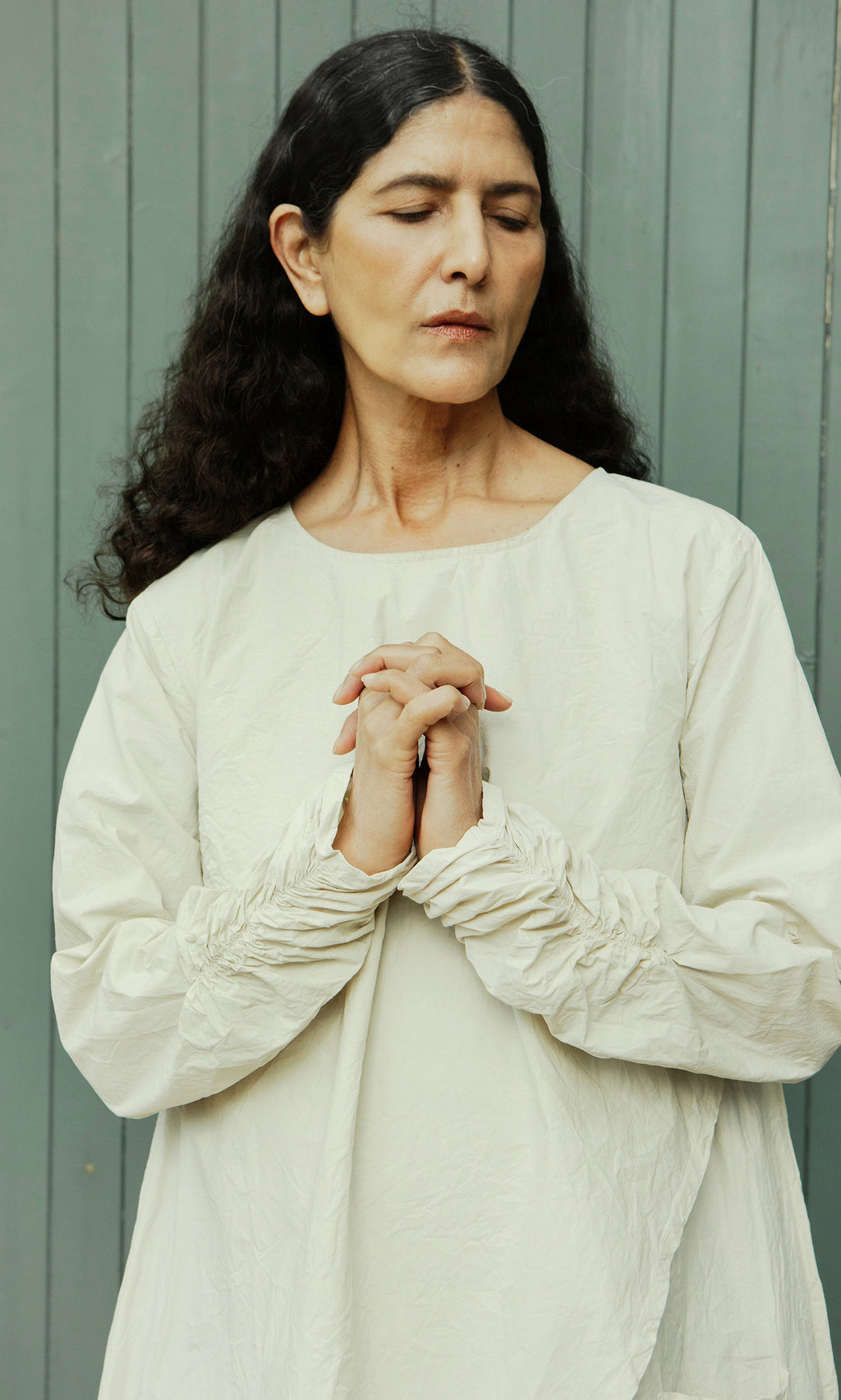 Plümo  Woman in a Clementine Blouse stands with hands clasped, eyes closed, against a gray wood backdrop.