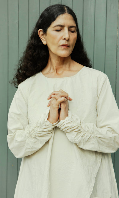 Plümo  Woman in a Clementine Blouse stands with hands clasped, eyes closed, against a gray wood backdrop.