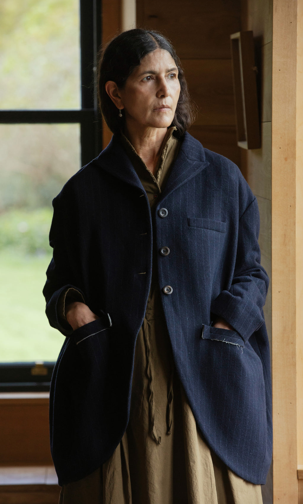 Plümo  Woman wears the Cleo Blazer in navy pinstripe, standing indoors by a window with hands in pockets, looking thoughtful.