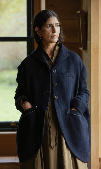 Plümo  Woman wears the Cleo Blazer in navy pinstripe, standing indoors by a window with hands in pockets, looking thoughtful.