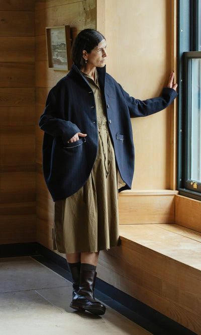 Plümo  Woman in the Cleo Blazer and a brown dress stands by a window, gazing outside with her hand on the sill.