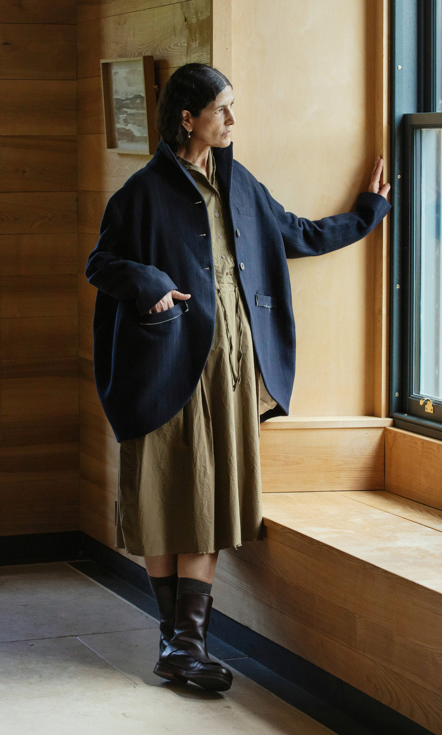 Plümo  Woman in the Cleo Blazer and a brown dress stands by a window, gazing outside with her hand on the sill.