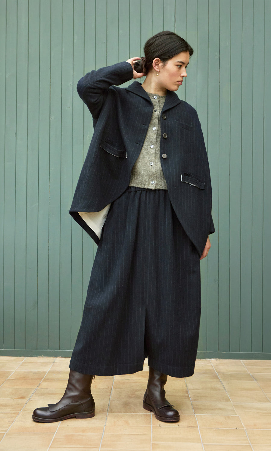 Plümo  Woman in the Cleo Blazer and boots stands by a green wall, channeling a vintage tailcoat silhouette.