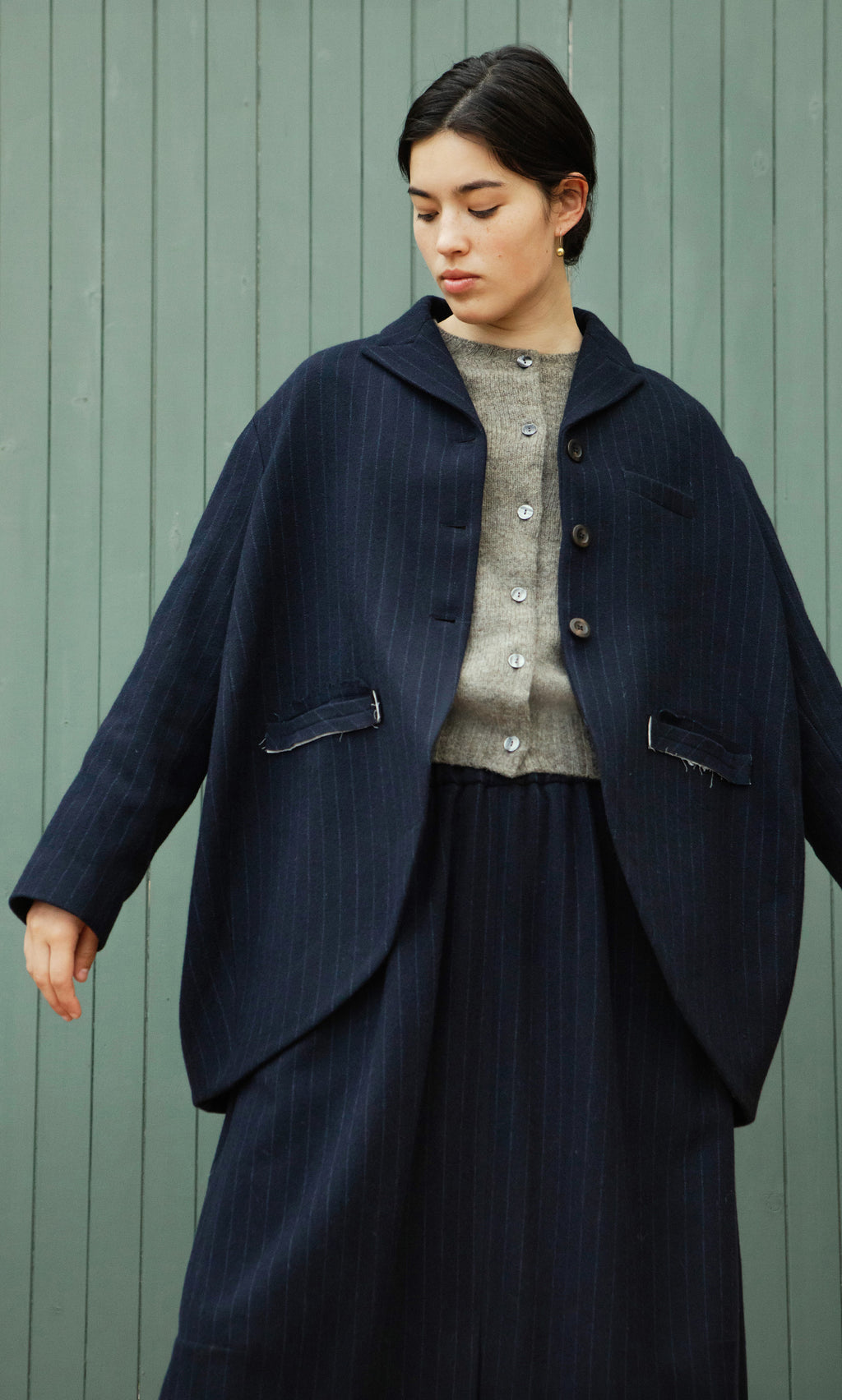 Plümo  Woman in Cleo Blazer and skirt over gray sweater stands before a light green wooden wall.