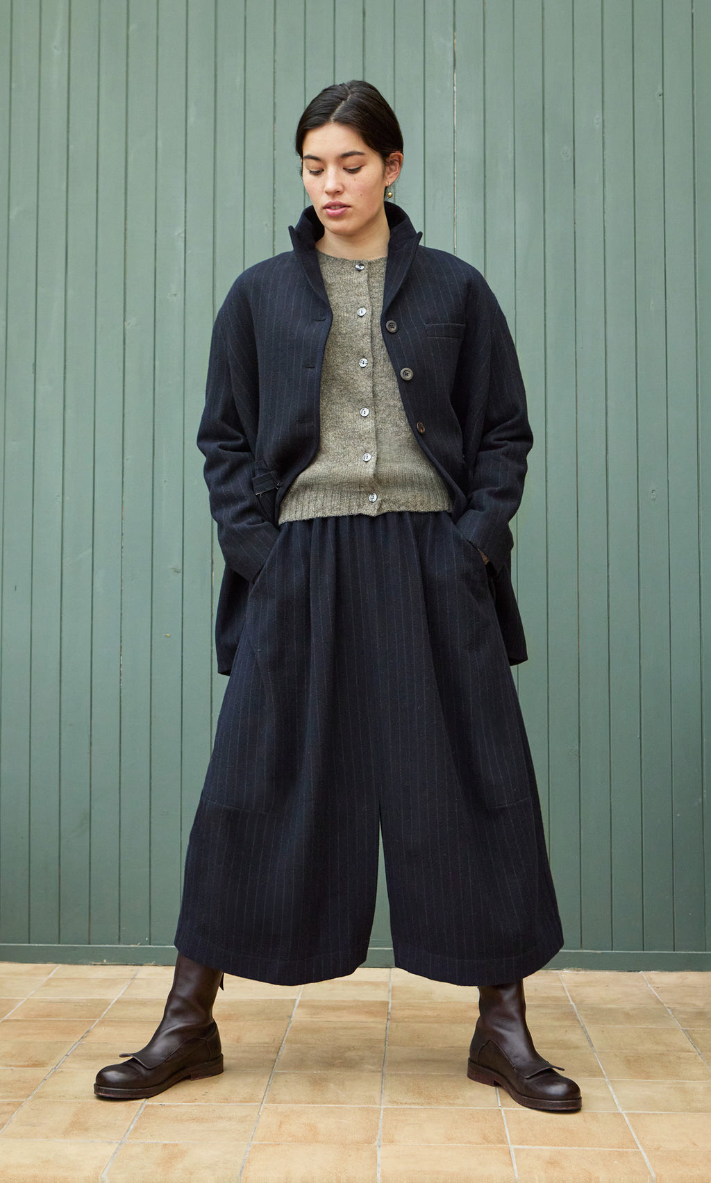 Plümo  Person in a gray buttoned sweater, black coat, boots, and Bo Skirt stands by a green paneled wall.