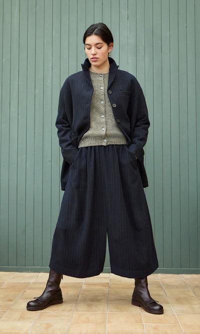 Plümo  Person in a gray buttoned sweater, black coat, boots, and Bo Skirt stands by a green paneled wall.
