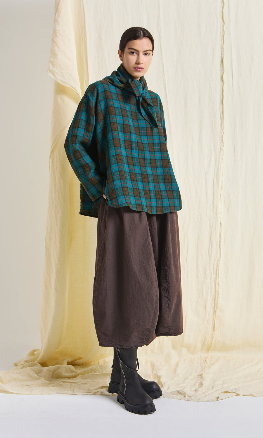 Plümo- Person wearing the Clodagh Blouse, wide brown pants, and black boots, standing before a beige fabric backdrop.