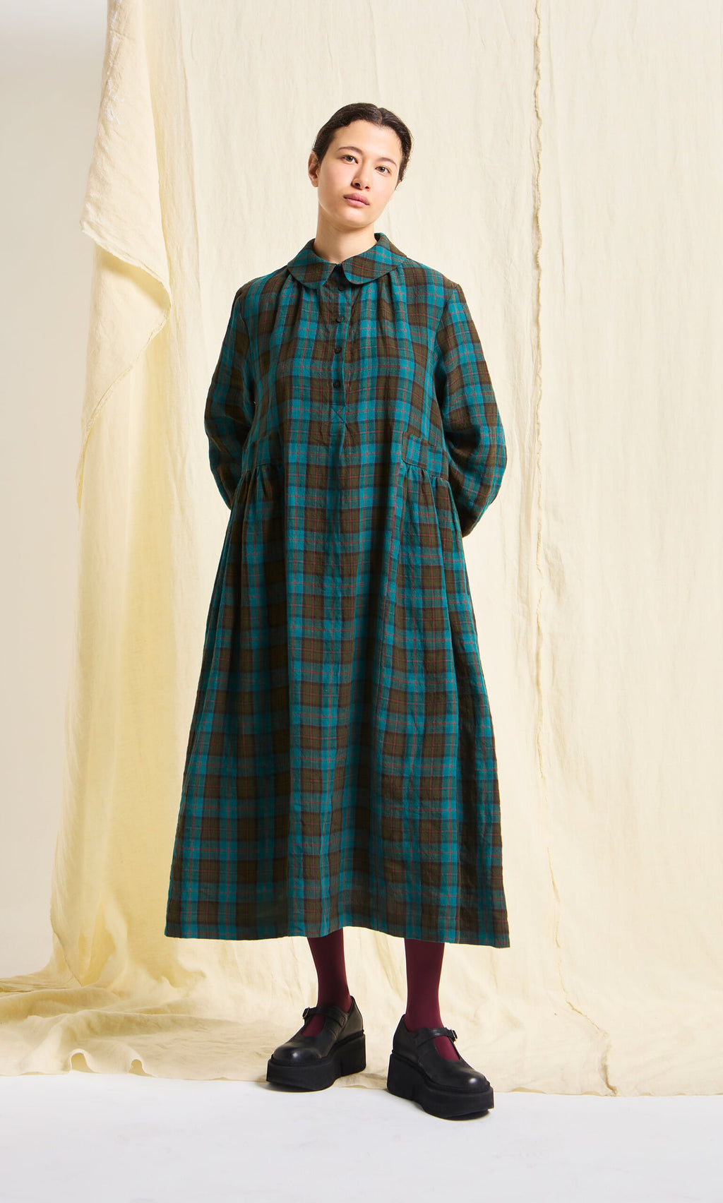 Plümo- A person stands before cream fabric wearing the Clodagh Tartan Dress with maroon tights and black shoes.