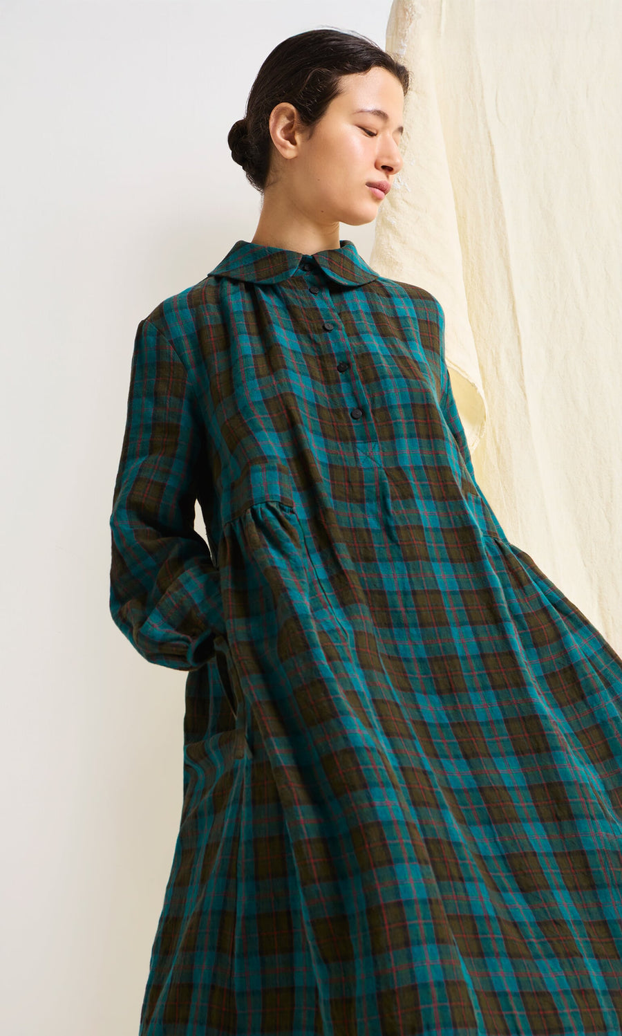 Plümo- Woman wearing the Clodagh Tartan Dress stands against a light background, looking to the side with hands in pockets.