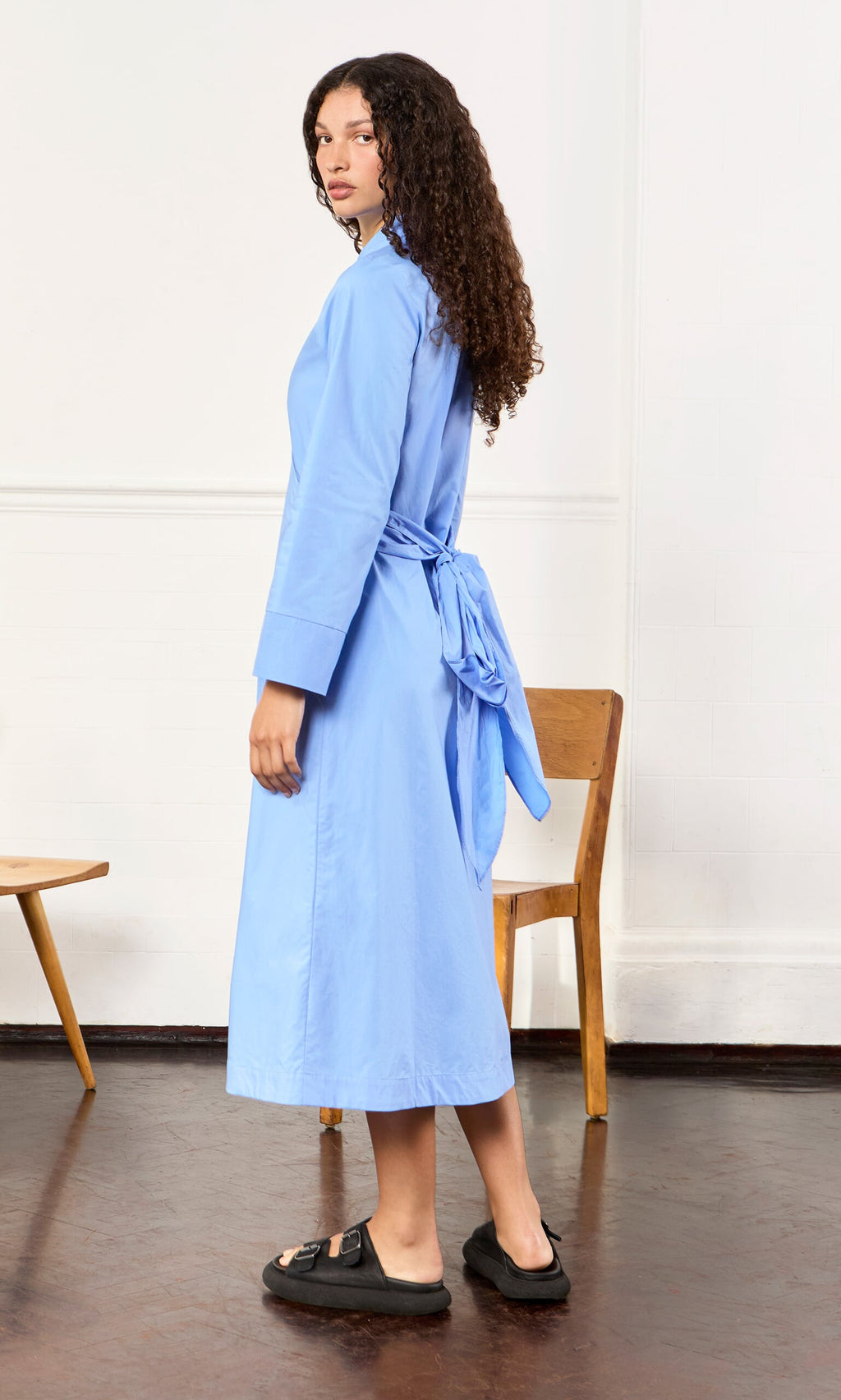 Plümo- Woman in the Cornflower Dress, a light blue cotton poplin shirt dress with tie belt, and black sandals near wooden chairs.
