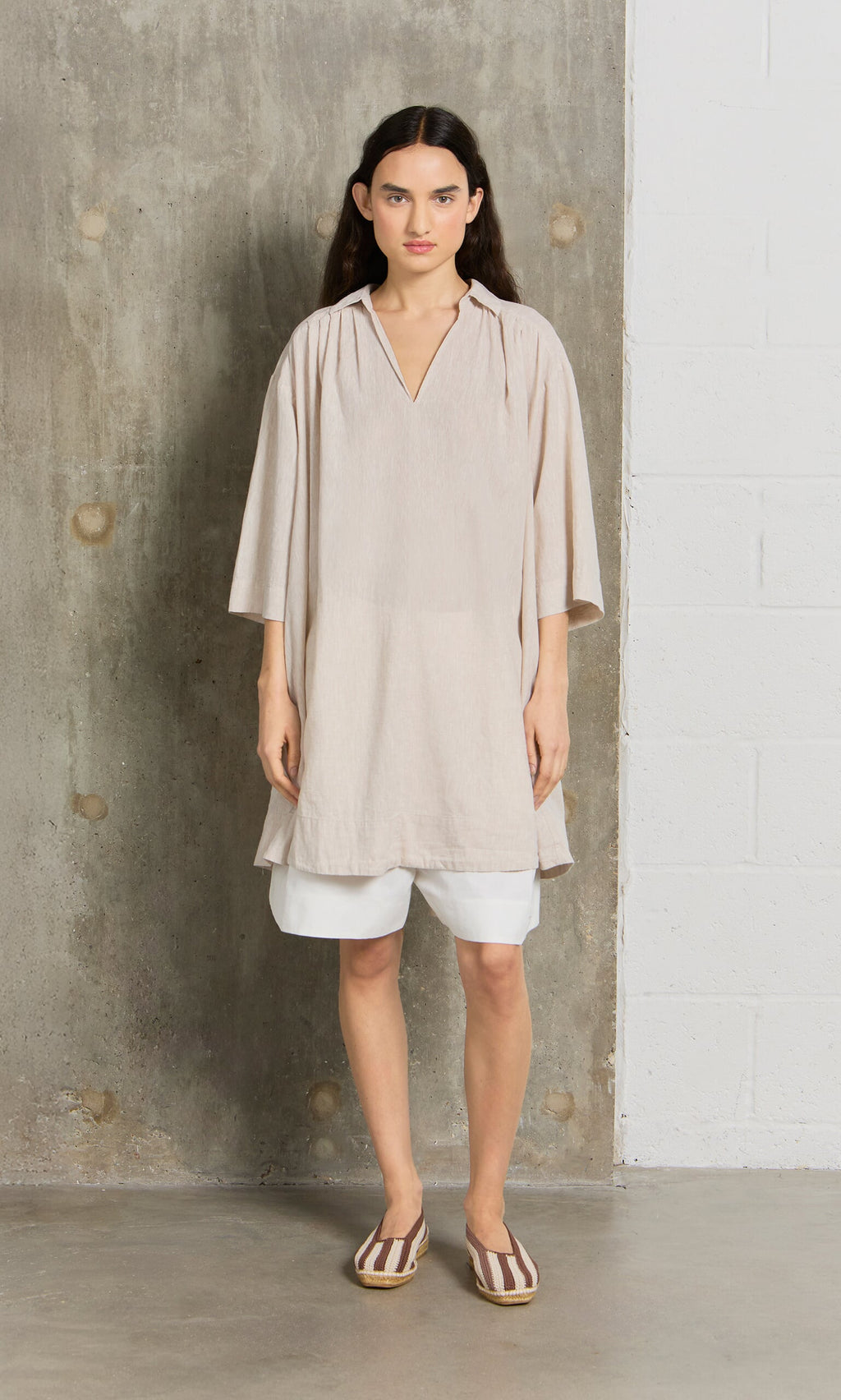 Plümo- Woman wears the Cory Tunic over white shorts and striped flats, standing by a concrete and white brick wall.