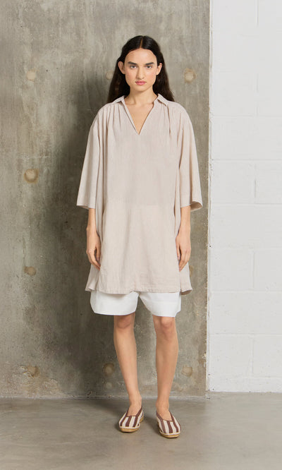 Plümo- Woman wears the Cory Tunic over white shorts and striped flats, standing by a concrete and white brick wall.