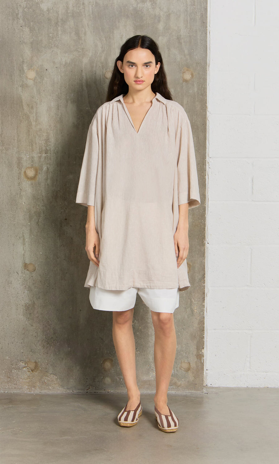 Plümo- Woman wears the Cory Tunic over white shorts and striped flats, standing by a concrete and white brick wall.