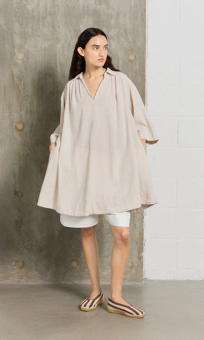 Plümo- Woman in the Cory Tunic over white shorts and striped espadrilles, standing against a concrete wall.