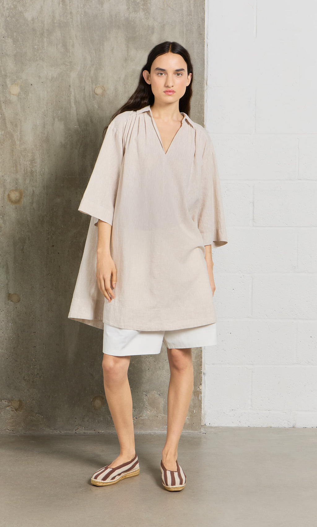 Plümo- A woman in a beige Cory Tunic, white shorts, and striped slip-ons stands by a concrete and white brick wall.
