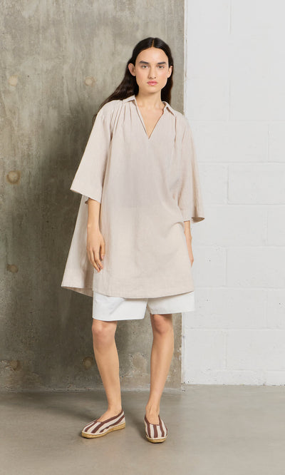 Plümo- A woman in a beige Cory Tunic, white shorts, and striped slip-ons stands by a concrete and white brick wall.