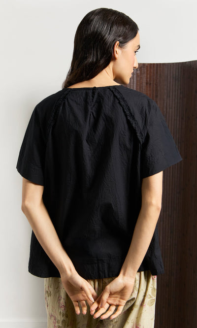 Plümo- A woman with dark hair wears the Crinkly Black Blouse, standing with her hands behind her back.