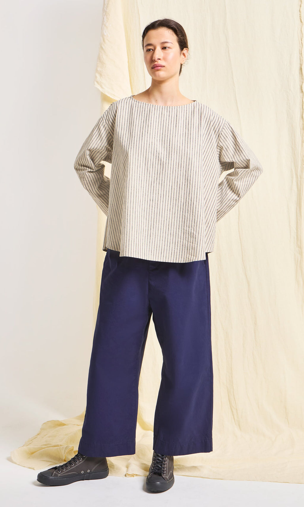 Plümo- Person wears a beige striped top, Cromer Pants - Navy, and black shoes, standing before a cream fabric backdrop.