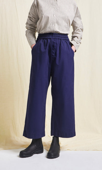 Plümo- Person in a striped shirt, Cromer Pants - Navy, and black boots stands against a light cream background.