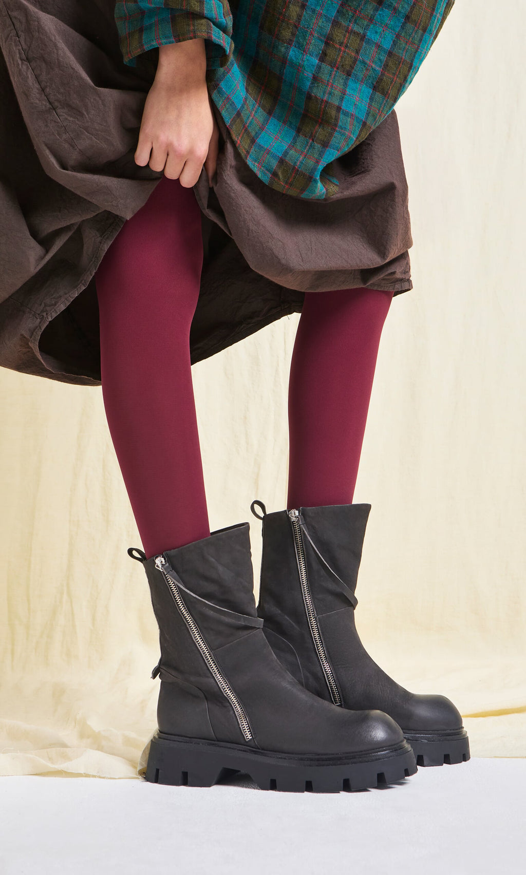 Plümo- Person wearing Deep Red Tights from Sweden and black zipper boots, holding up a brown and green plaid skirt.