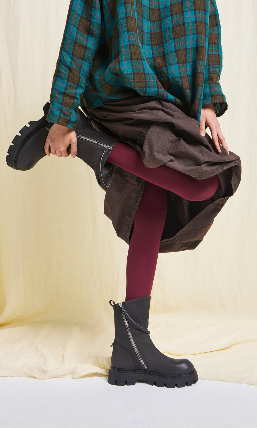 Plümo- A person in a plaid shirt and Deep Red Tights from Sweden lifts one leg to adjust their black boots.