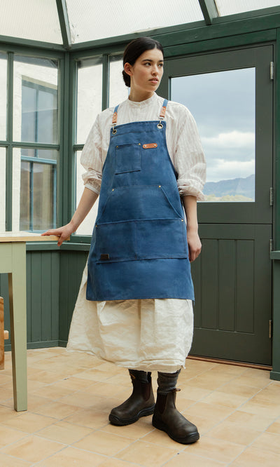 Plümo  Woman in a white dress and Denim Cobbler Apron stands by a table indoors, green windows in the background.