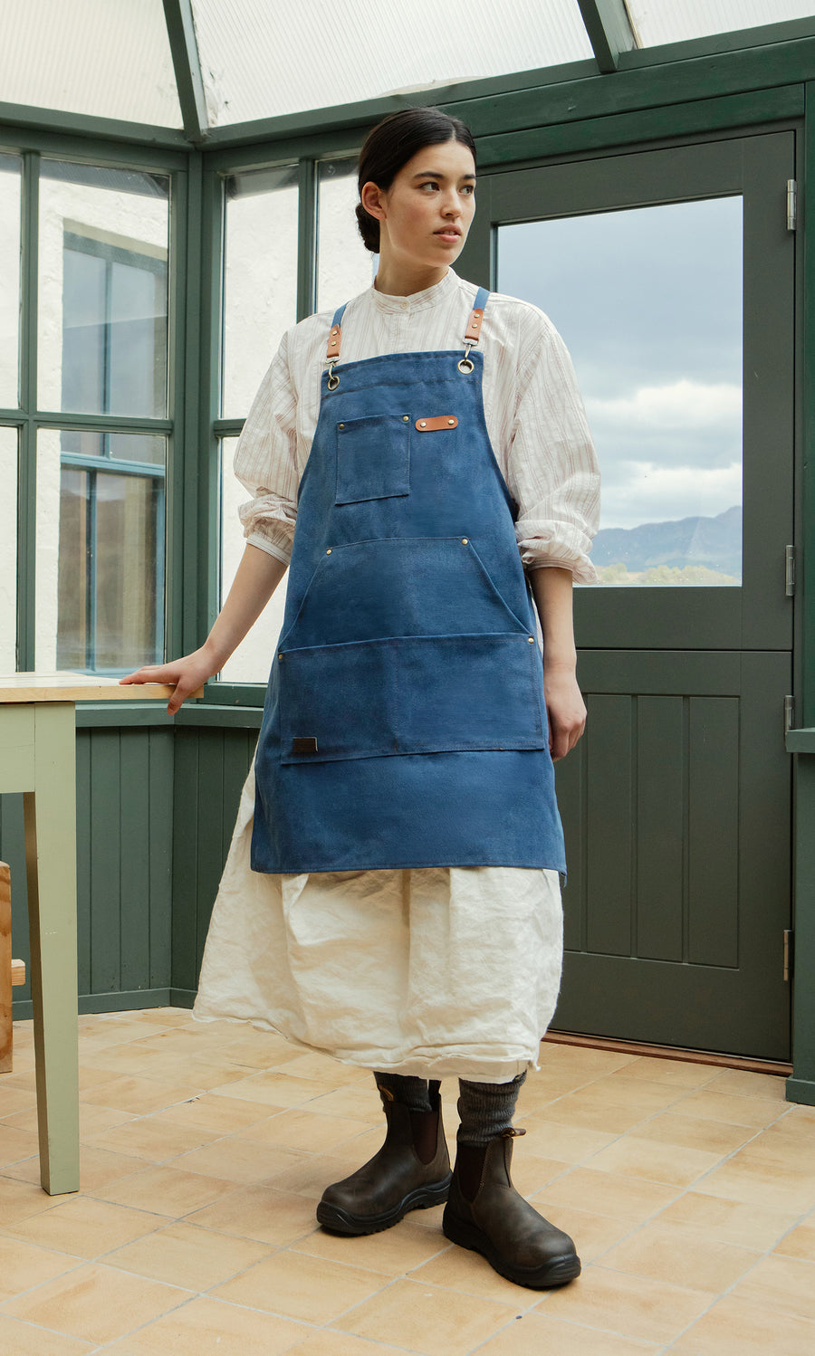 Plümo  Woman in a white dress and Denim Cobbler Apron stands by a table indoors, green windows in the background.