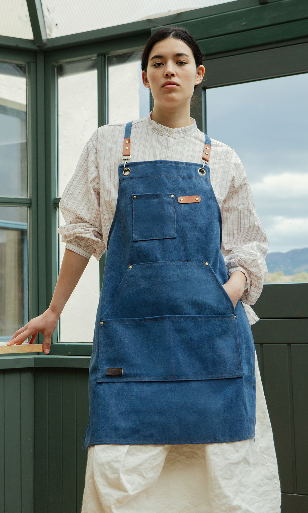 Plümo  Person wears a Denim Cobbler Apron with utility pockets over a striped shirt, standing by a window with a neutral expression.