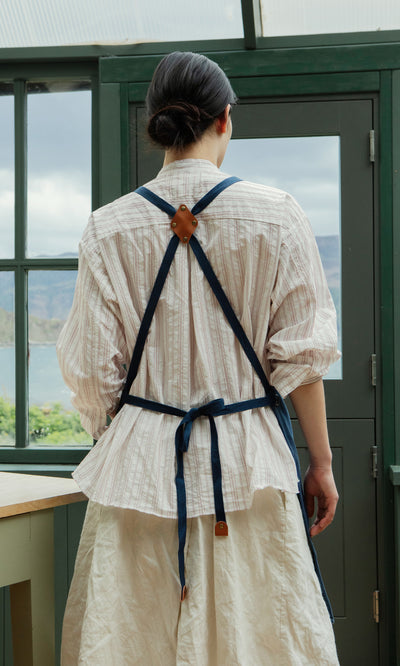 Plümo  Person with dark hair in a bun wears a striped shirt and Denim Cobbler Apron, standing in a bright room.