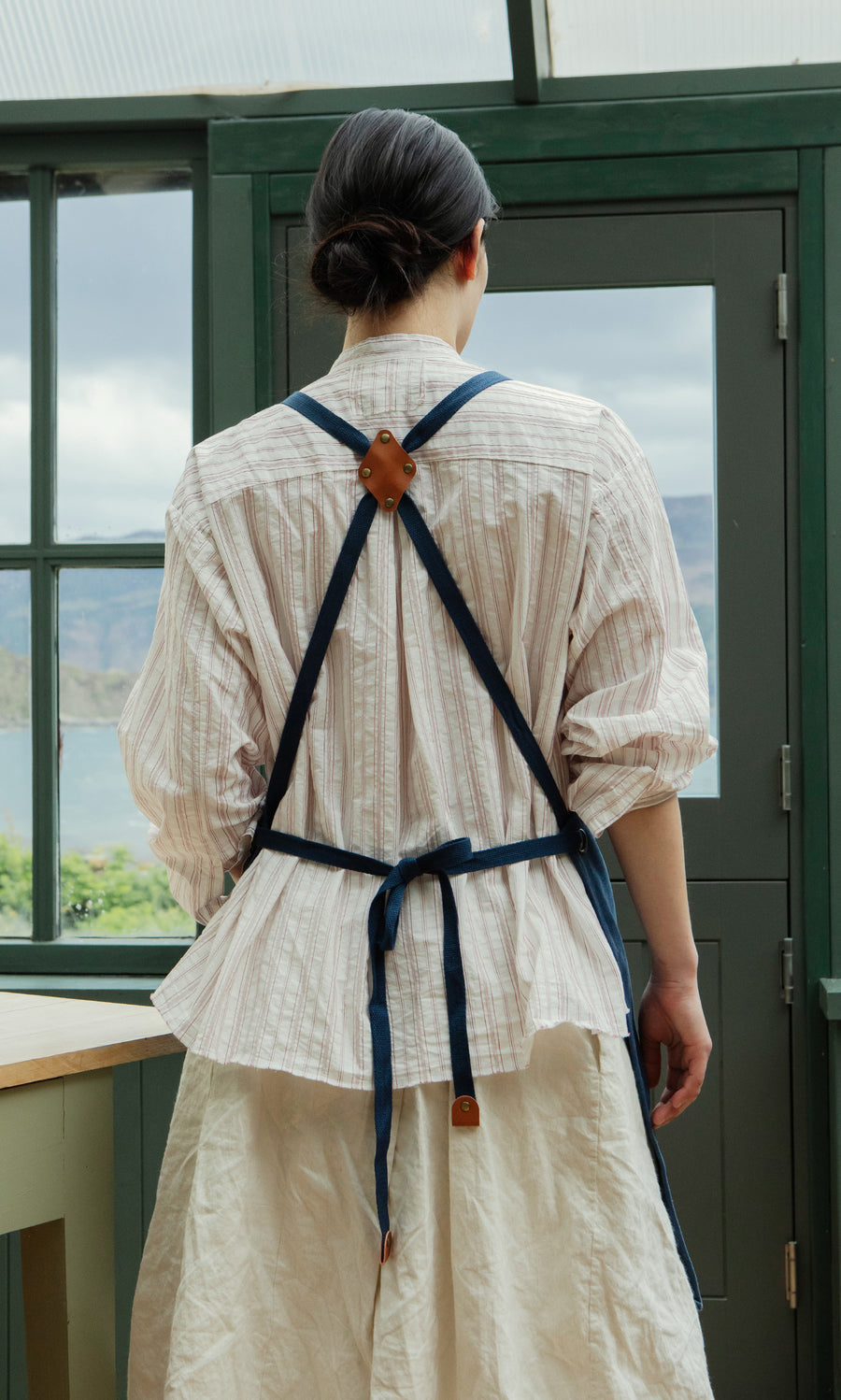 Plümo  Person with dark hair in a bun wears a striped shirt and Denim Cobbler Apron, standing in a bright room.