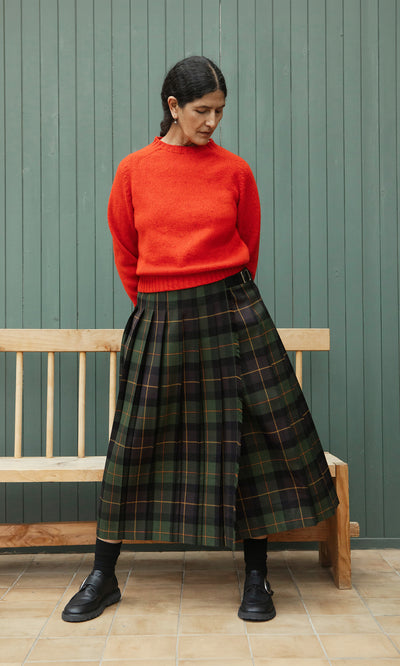 Plümo  Woman in a red sweater and Derry Kilt stands by a green wall and bench, looking down.
