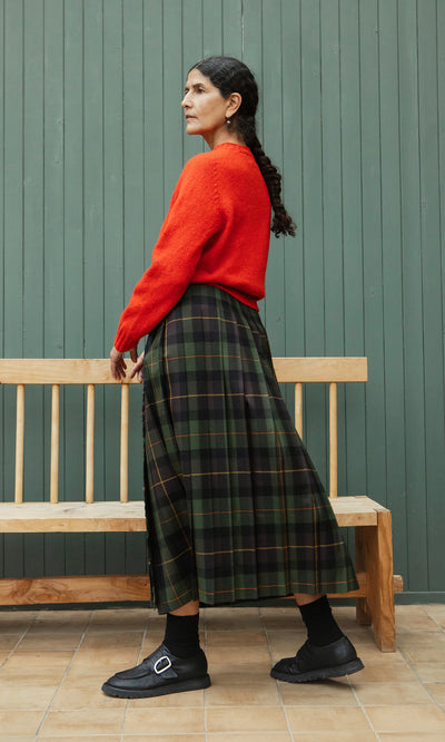 Plümo  Woman in red sweater and Derry Kilt stands by wooden bench, green paneled wall behind her.