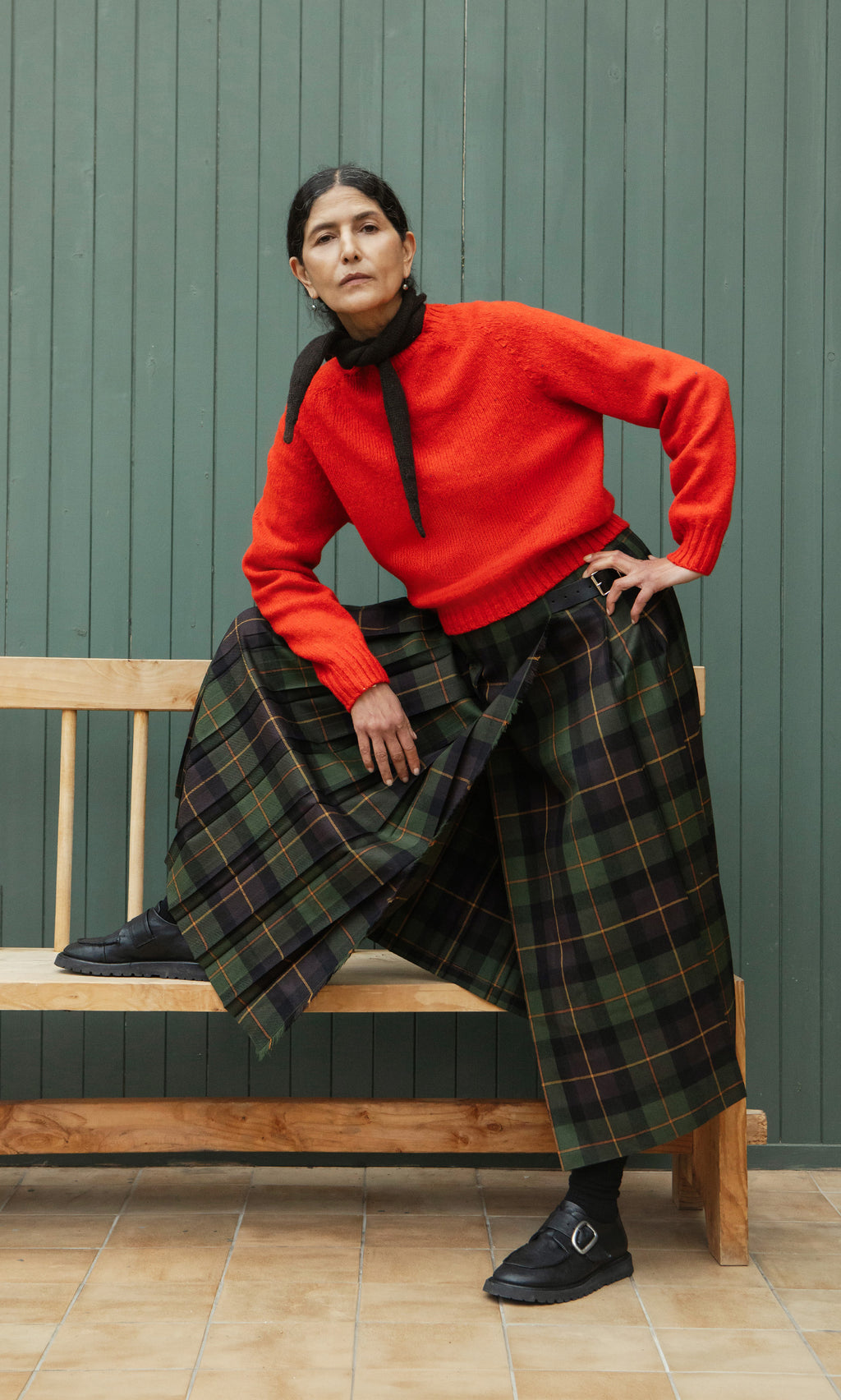 Plümo  A woman in a red sweater and Derry Kilt poses on a wooden bench against a green paneled wall.