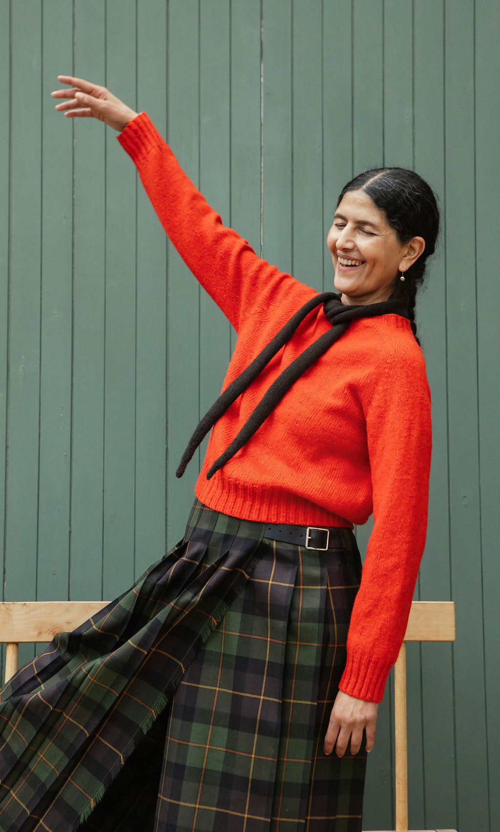 Plümo  Woman in red sweater and Derry Kilt smiles with eyes closed, posing by a green wall.