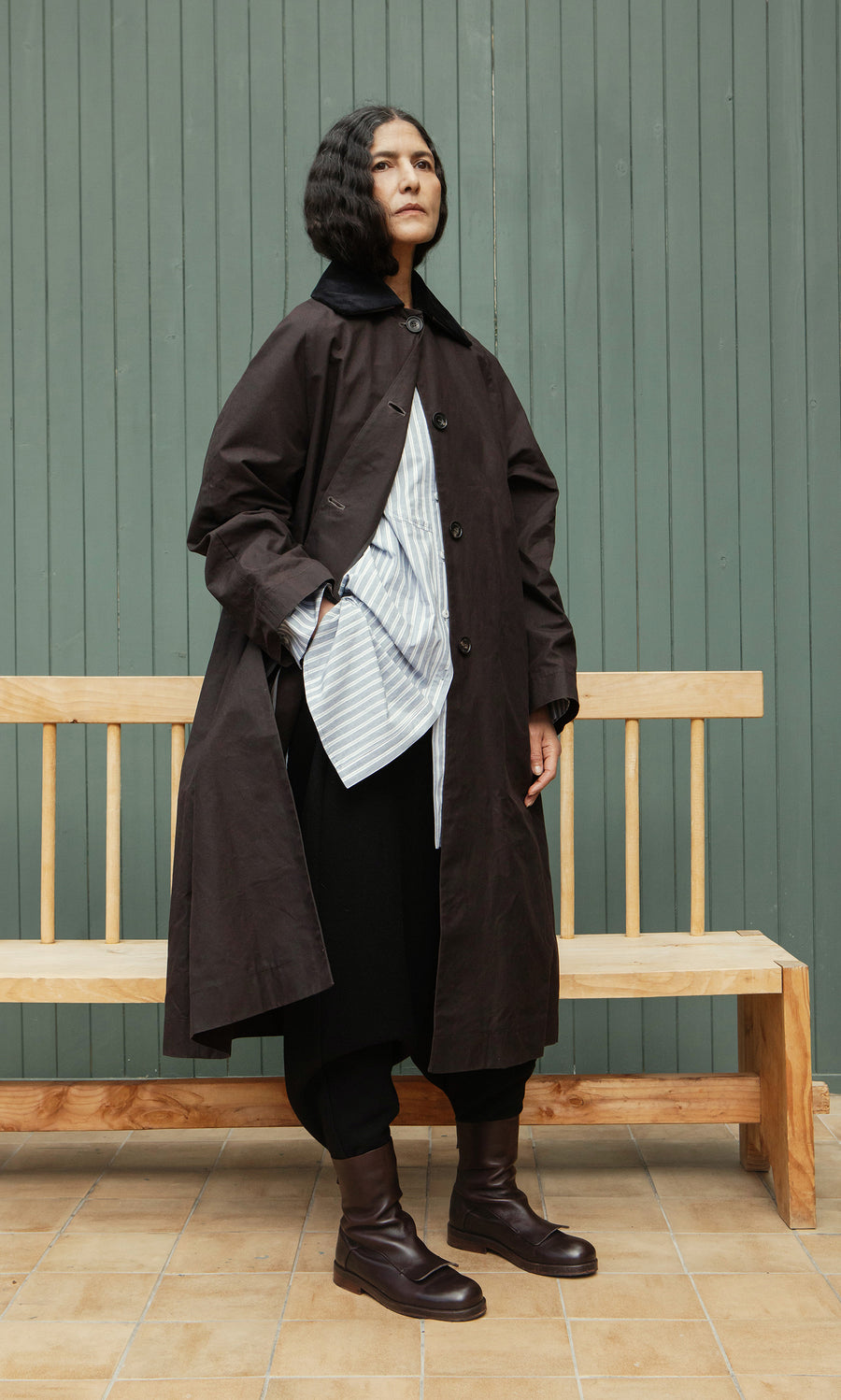 Plümo  A person with dark wavy hair wears the Doherty Coat - Soil over a striped shirt, leaning on a wooden bench.