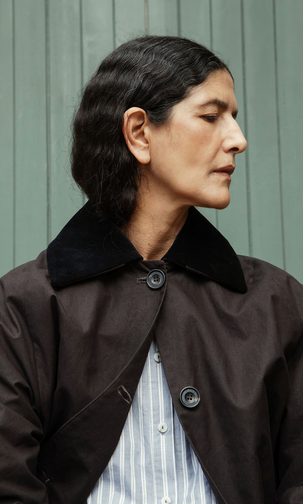 Plümo  Person with dark hair in the Doherty Coat - Soil and a striped shirt, looking to the side before a green wall.
