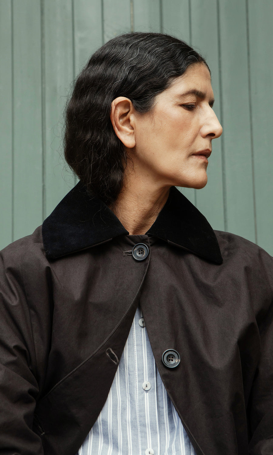 Plümo  Person with dark hair in the Doherty Coat - Soil and a striped shirt, looking to the side before a green wall.