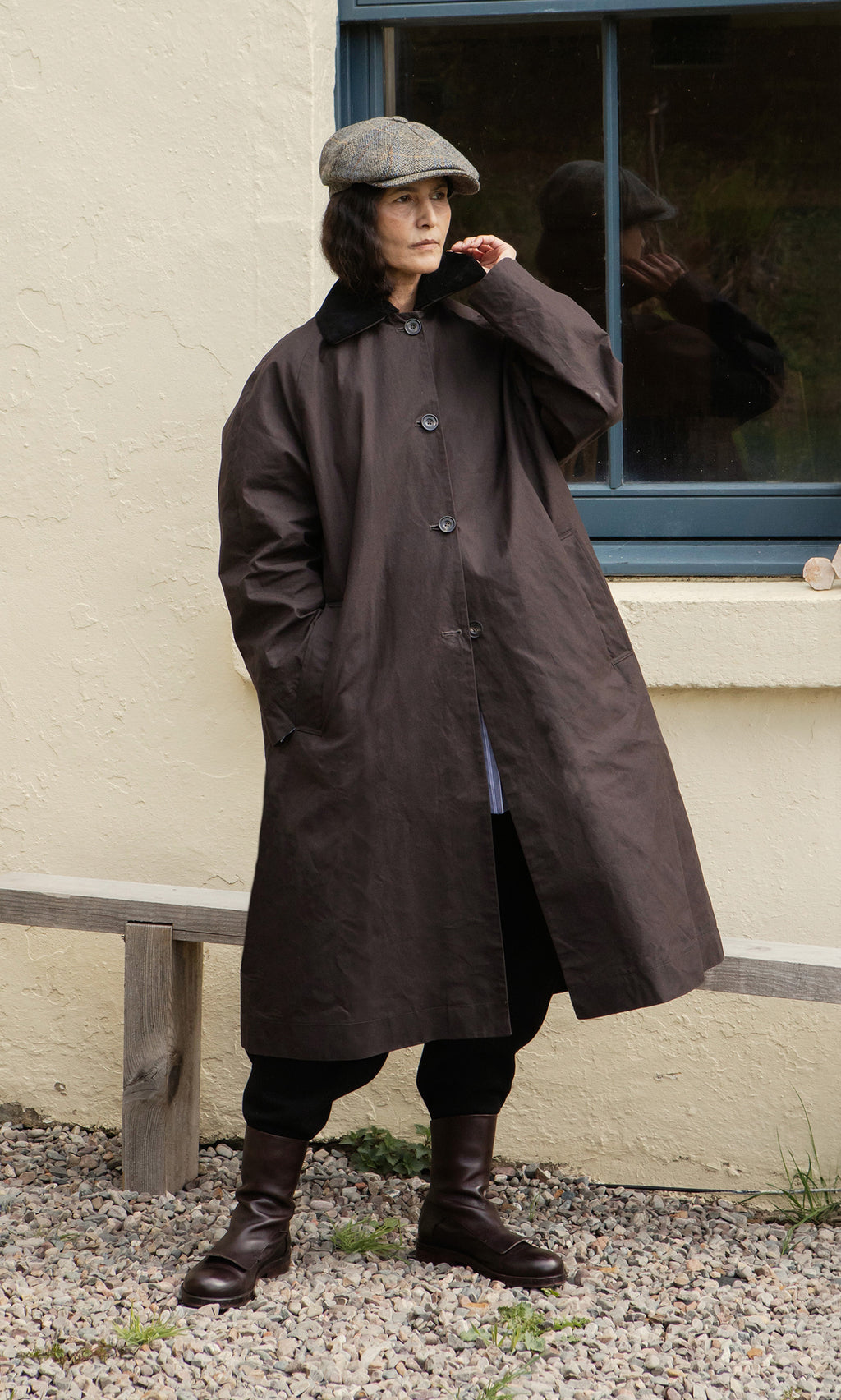 Plümo  Individual wearing the Doherty Coat - Soil, flat cap, and boots stands by a window and bench on gravel outdoors.