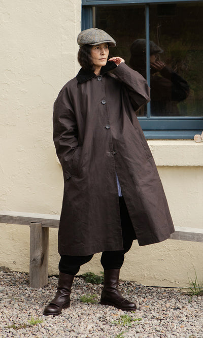 Plümo  Individual wearing the Doherty Coat - Soil, flat cap, and boots stands by a window and bench on gravel outdoors.