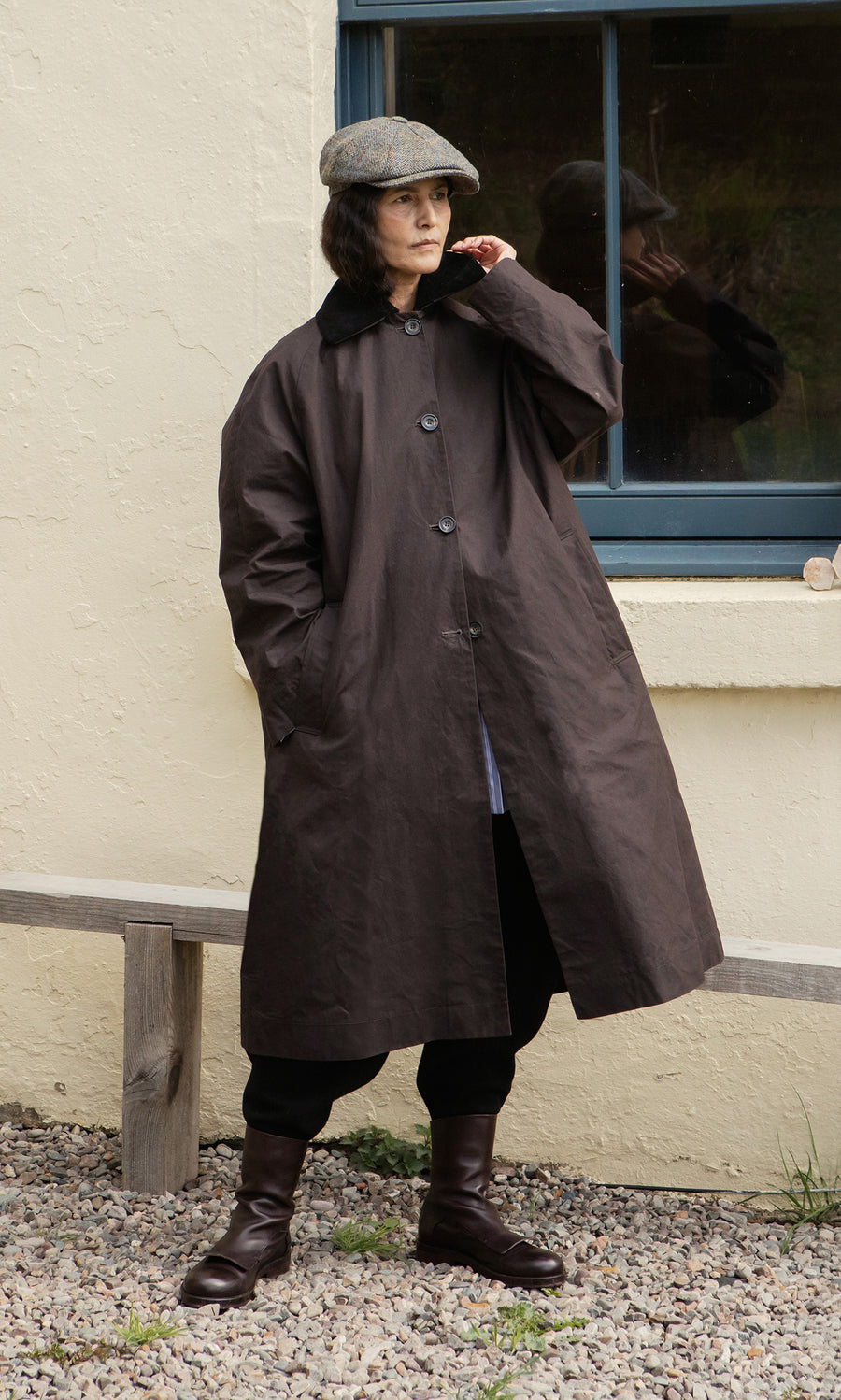 Plümo  Individual wearing the Doherty Coat - Soil, flat cap, and boots stands by a window and bench on gravel outdoors.
