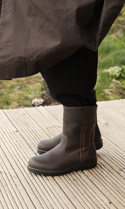 Plümo  Person in Rigger Boots with cushioned removable sole stands on a wooden deck; brown leather and stitching show.