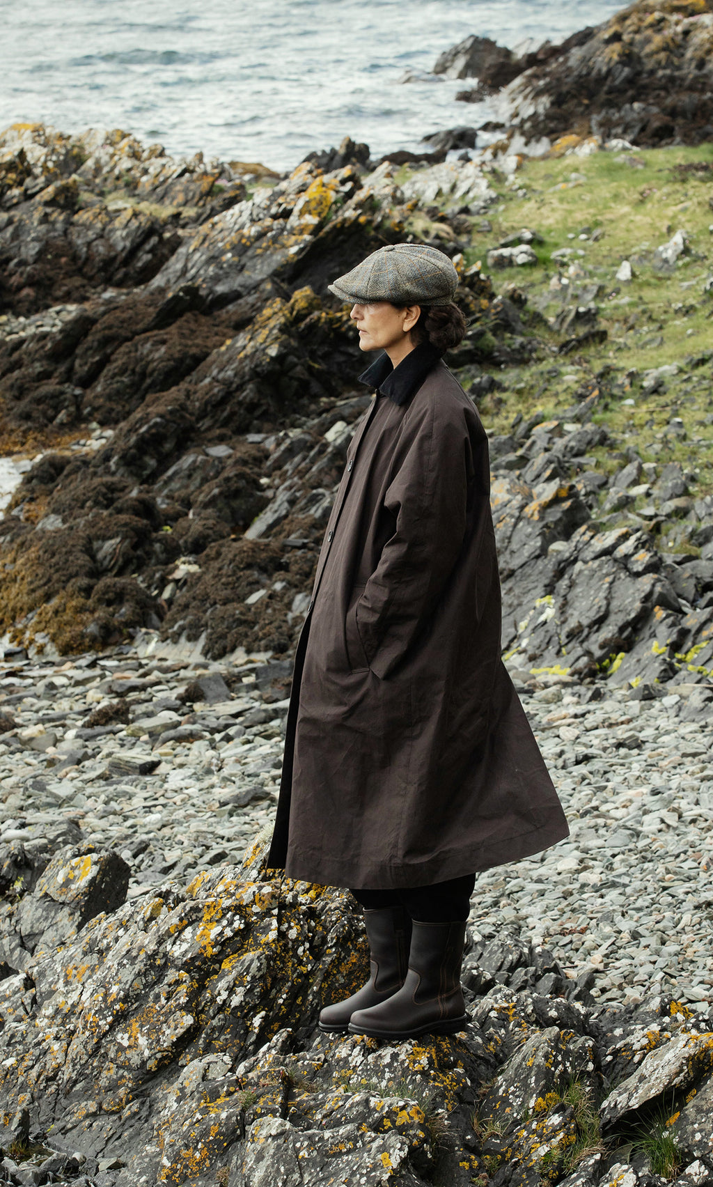 Plümo  Wearing the Doherty Coat - Soil and a cap, a figure stands on rocky shore, looking out at the sea.