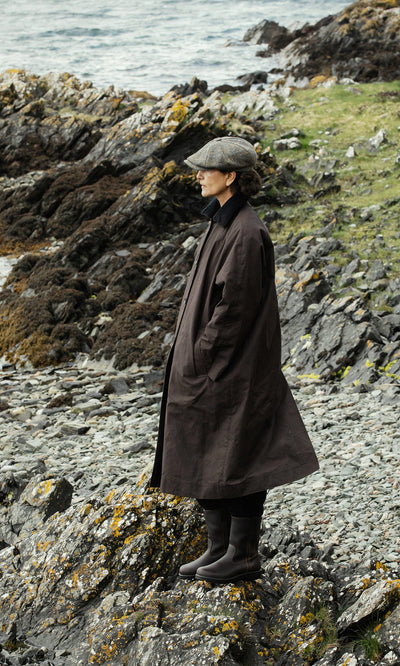 Plümo  Wearing the Doherty Coat - Soil and a cap, a figure stands on rocky shore, looking out at the sea.